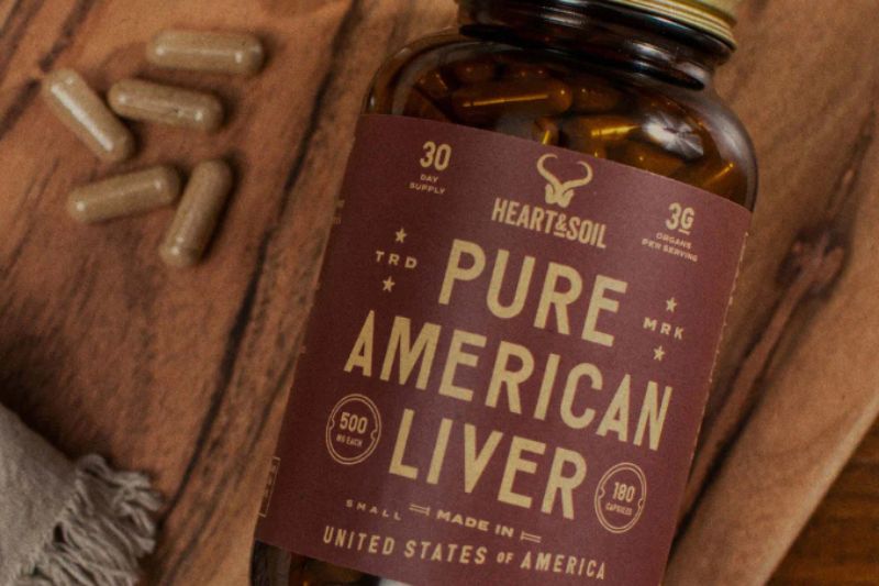 A glass bottle containing capsules with the label features a design with the words "Heart & Soil" and "Pure American Liver" in bold.