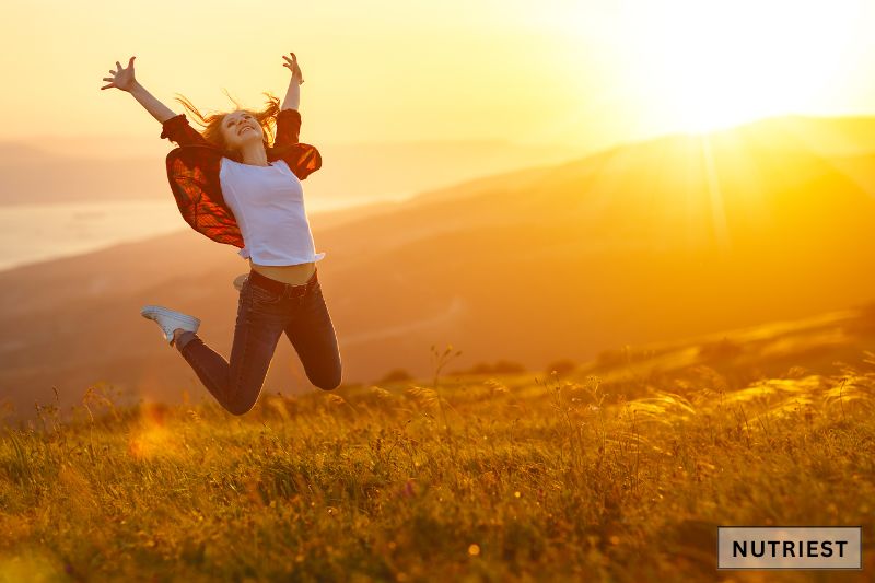 The image shows a young woman joyfully jumping in a sunlit field, with her arms raised high emphasizing the connection between natural supplements and enhanced vitality.