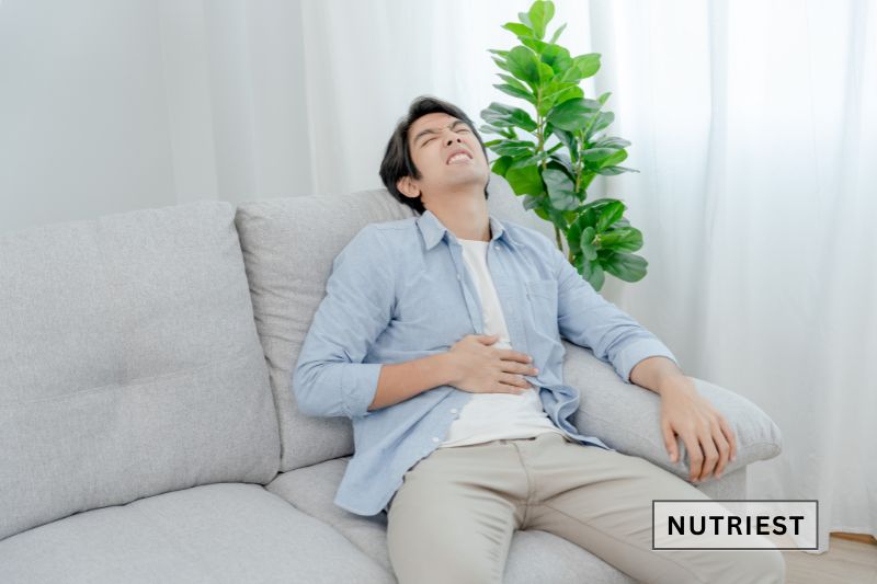 A man sitting on a couch, clutching his stomach with a pained expression, possibly experiencing digestive discomfort.