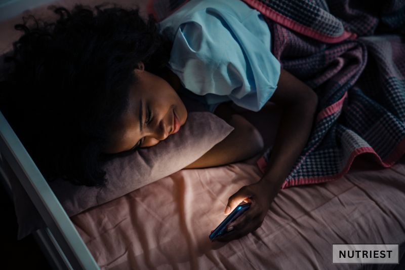 Woman using phone in bed at night, highlighting habits that may cause sallow skin.